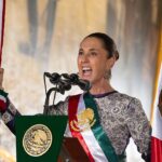 Claudia Sheinbaum habla ante micrófonos con la bandera de México al fondo
