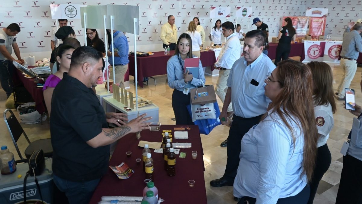 Participantes conversan junto a una mesa de productos en una exhibición de Hecho en Tamaulipas.